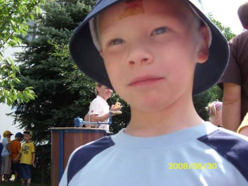Ein Junge mit einem blauen T-Shirt und einem Hut steht im Freien, im Hintergrund weitere Kinder.